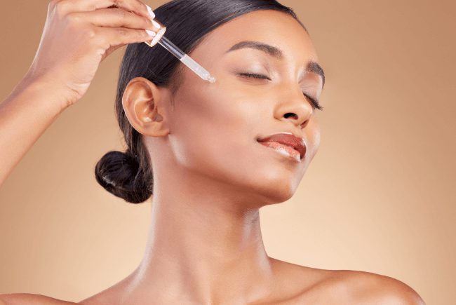 Femme avec une peau lumineuse qui applique une goutte de sérum sur son visage les yeux fermés.